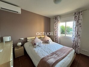 A bedroom with a bed, a nightstand, an air conditioner, a window with curtains, and a view of greenery outside. - Detached house in Calonge with private pool and parking (Calonge)