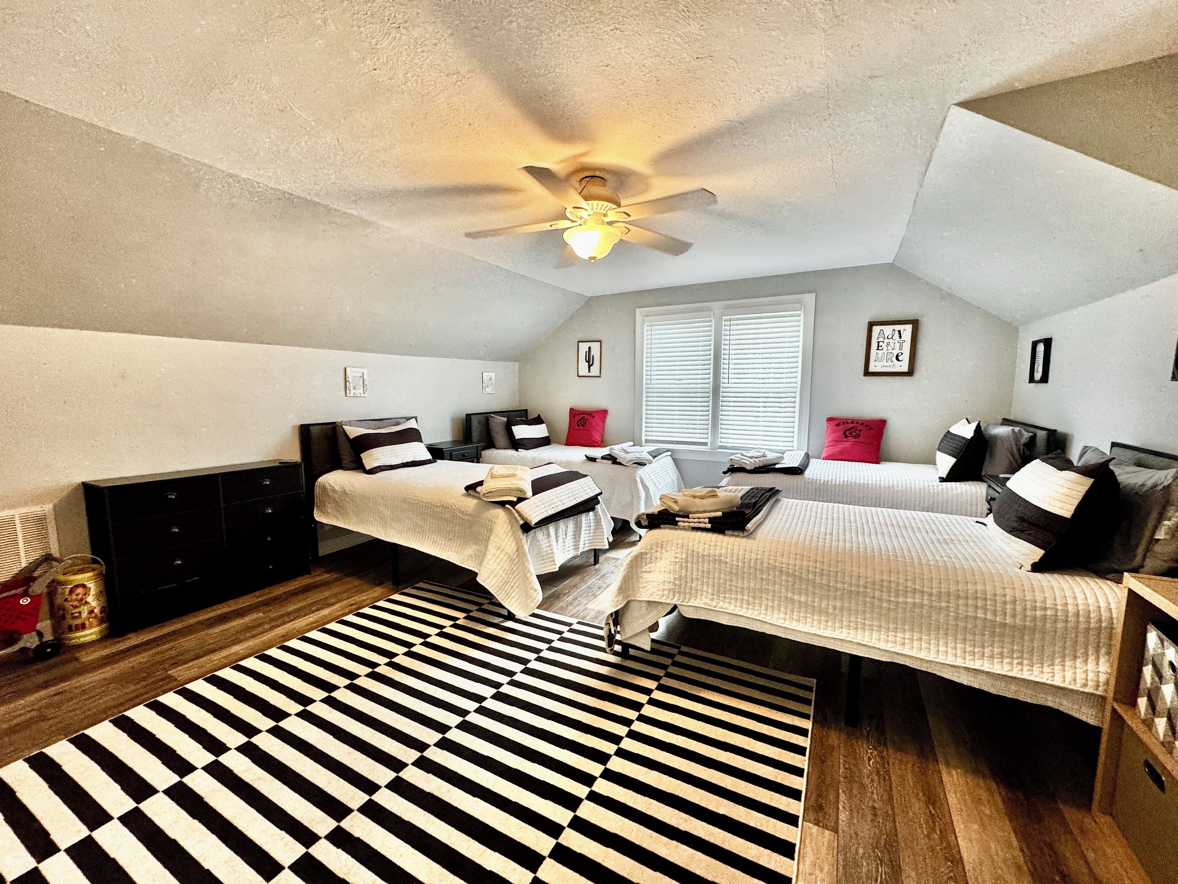 A bedroom with two beds, a ceiling fan, a window with blinds, and a black and white striped rug.