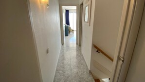 Un couloir étroit, avec un luminaire sur le côté gauche, une porte sur le côté droit, et, au loin, on aperçoit un salon.