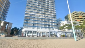 A high-rise hotel with multiple floors, a ground-level entrance, and a beach in the foreground.