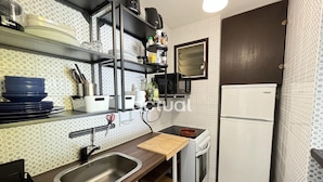 A compact kitchen with a sink, a stove, and a refrigerator. There are shelves above the sink holding dishes and kitchen items.