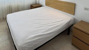 Una cama individual con cabecera de madera, una mesita de noche y un aparador de madera en una habitación con suelo de baldosas y cortinas translúcidas.