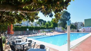 Una piscina con sillas de descanso y mesas, rodeada por una valla y follaje.