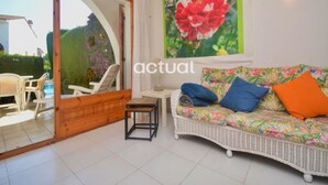 A room with a white wicker sofa, a small table, and a chair. There is a large window with a view of a garden and a pool. - Cosy townhouse, near beach, terrace, communal pool, WiFi and parking in Platja d'Aro (Platja d'Aro)
