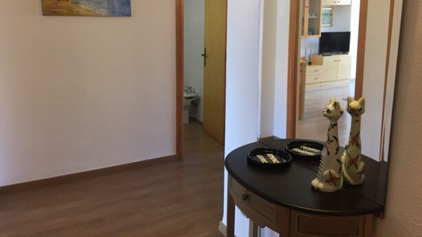 A hallway with a wooden floor, a painting on the wall, a mirror reflecting a kitchen, and a small table with decorative items. - 3-bedroom apartment, pool & parking, 350 m from the beach (Platja d'Aro)