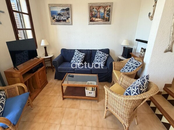 Una sala de estar con sofá, una mesa de centro y sillones de mimbre. Hay un televisor sobre un mueble de madera y dos cuadros en la pared.