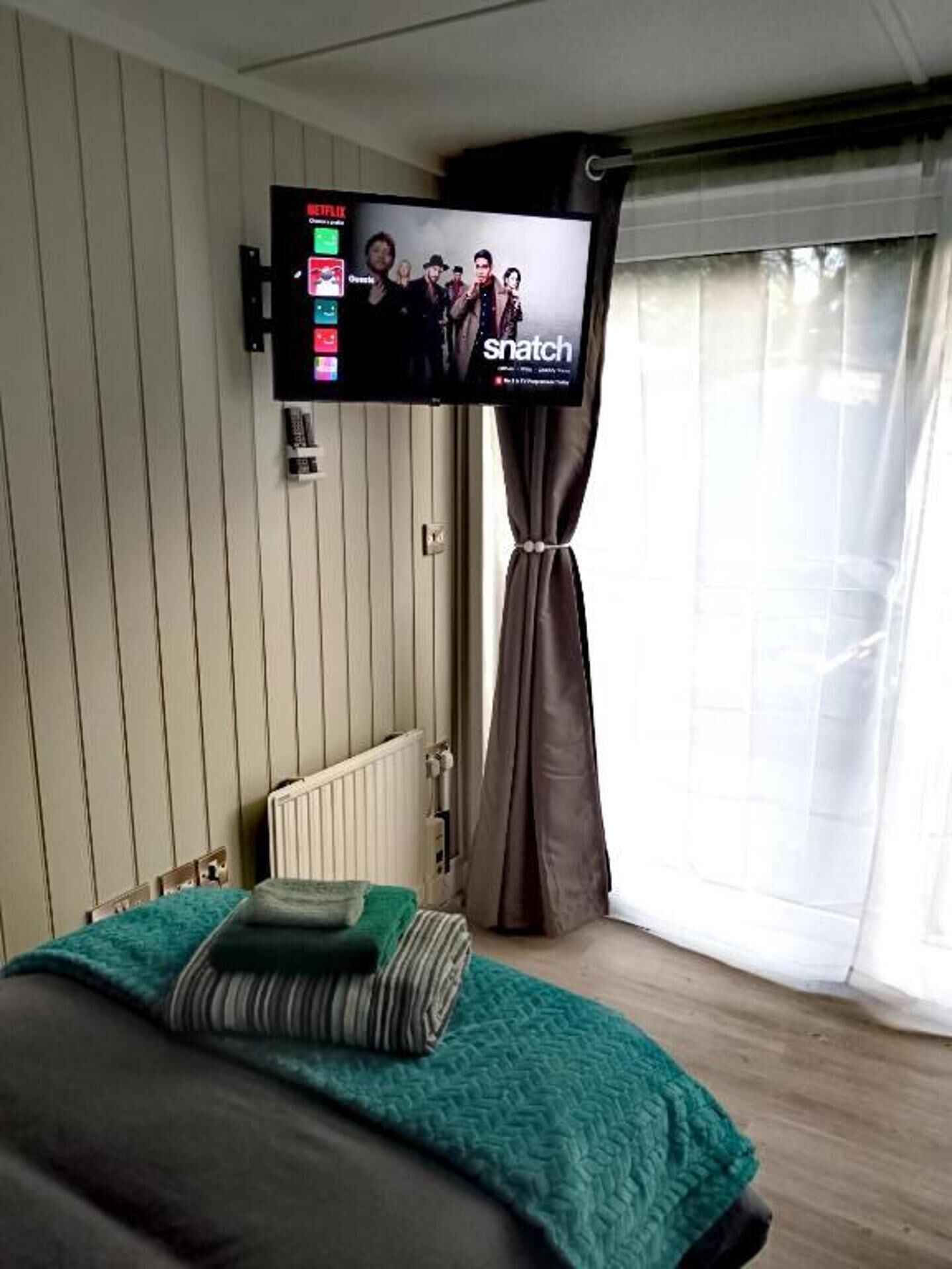 Ένα υπνοδωμάτιο με ένα κρεβάτι, μια τηλεόραση τοποθετημένη στον τοίχο, ένα καλοριφέρ και ένα παράθυρο με κουρτίνες.