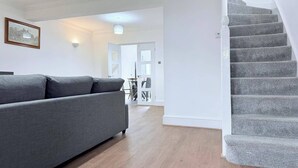A modern living room with a grey sofa, wooden flooring, a staircase, and a dining area in the background.