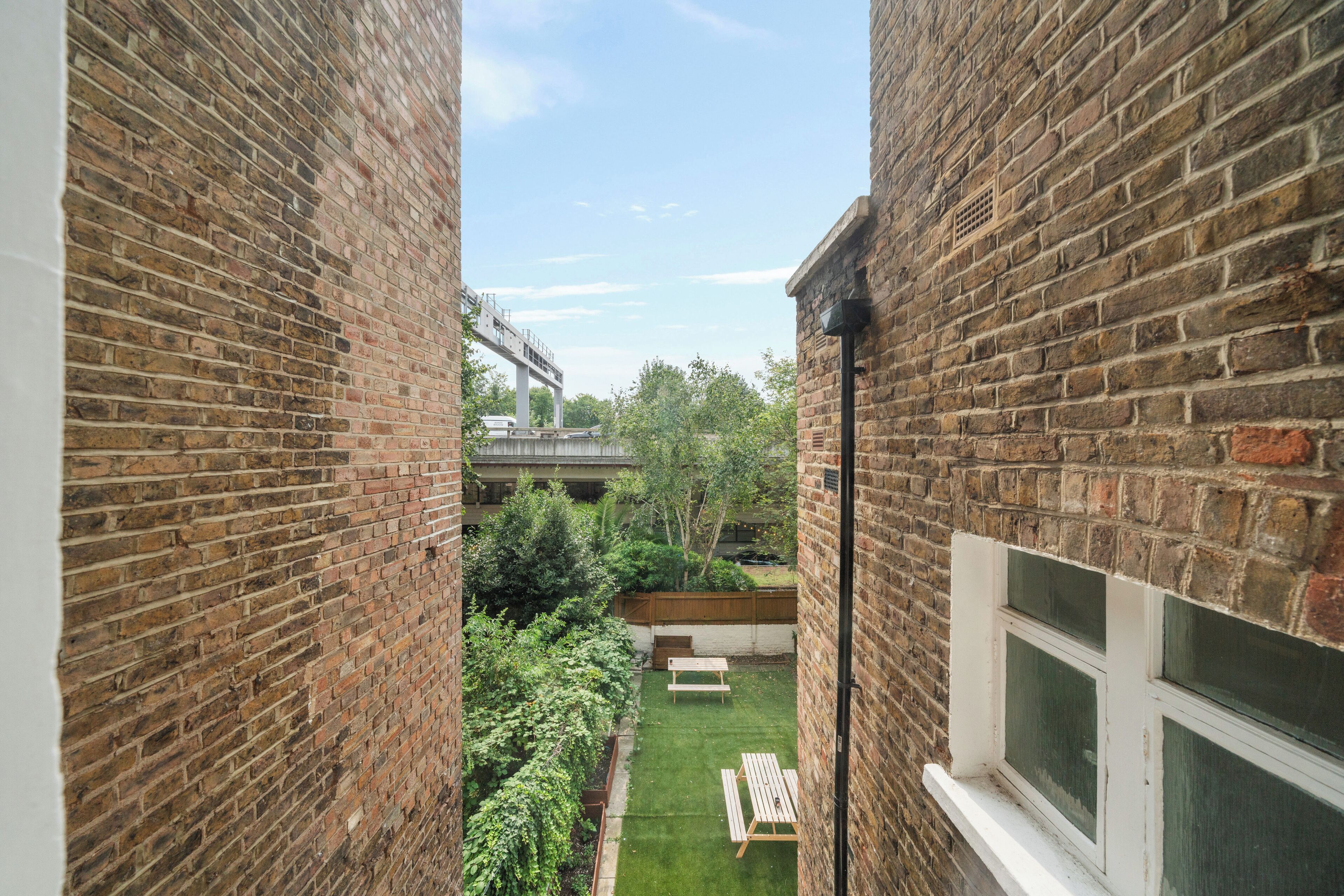 A narrow brick corridor with a small garden area, a wooden bench, and a window.