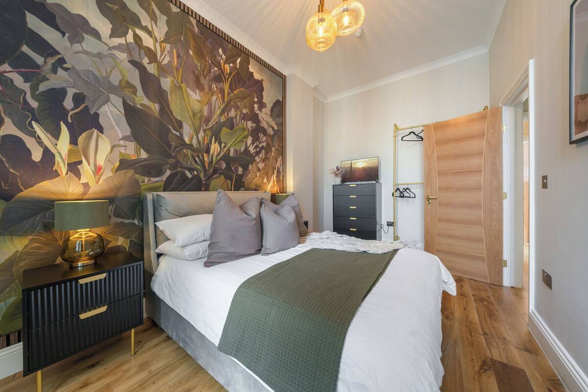 A bedroom with a large floral mural, a bed with a white comforter and green throw, a nightstand with a lamp, and a wooden door.