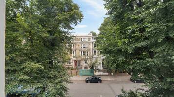 A street view with a building that has a classic architectural style, surrounded by lush greenery and parked cars.