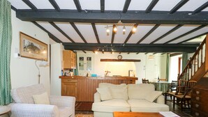 A cozy living room with a wooden ceiling, a sofa, a chair, a coffee table, and a kitchen area in the background.