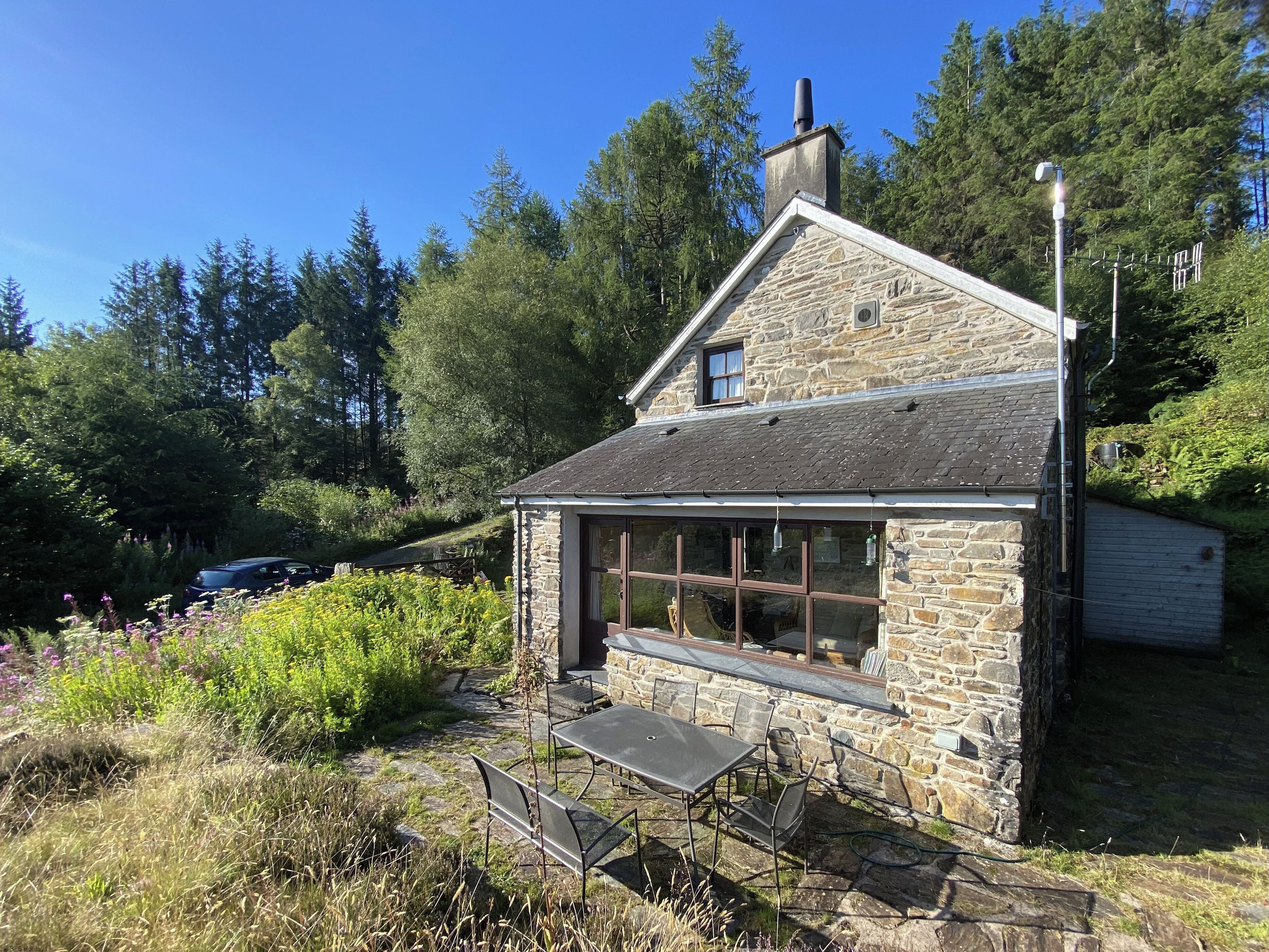A stone cottage with a large window, a chimney, and a garden area with a table and chairs.