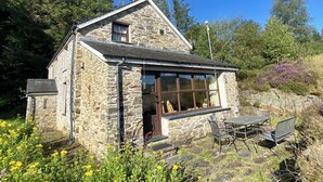 A stone cottage with a small patio, a chimney, and a window overlooking a garden.