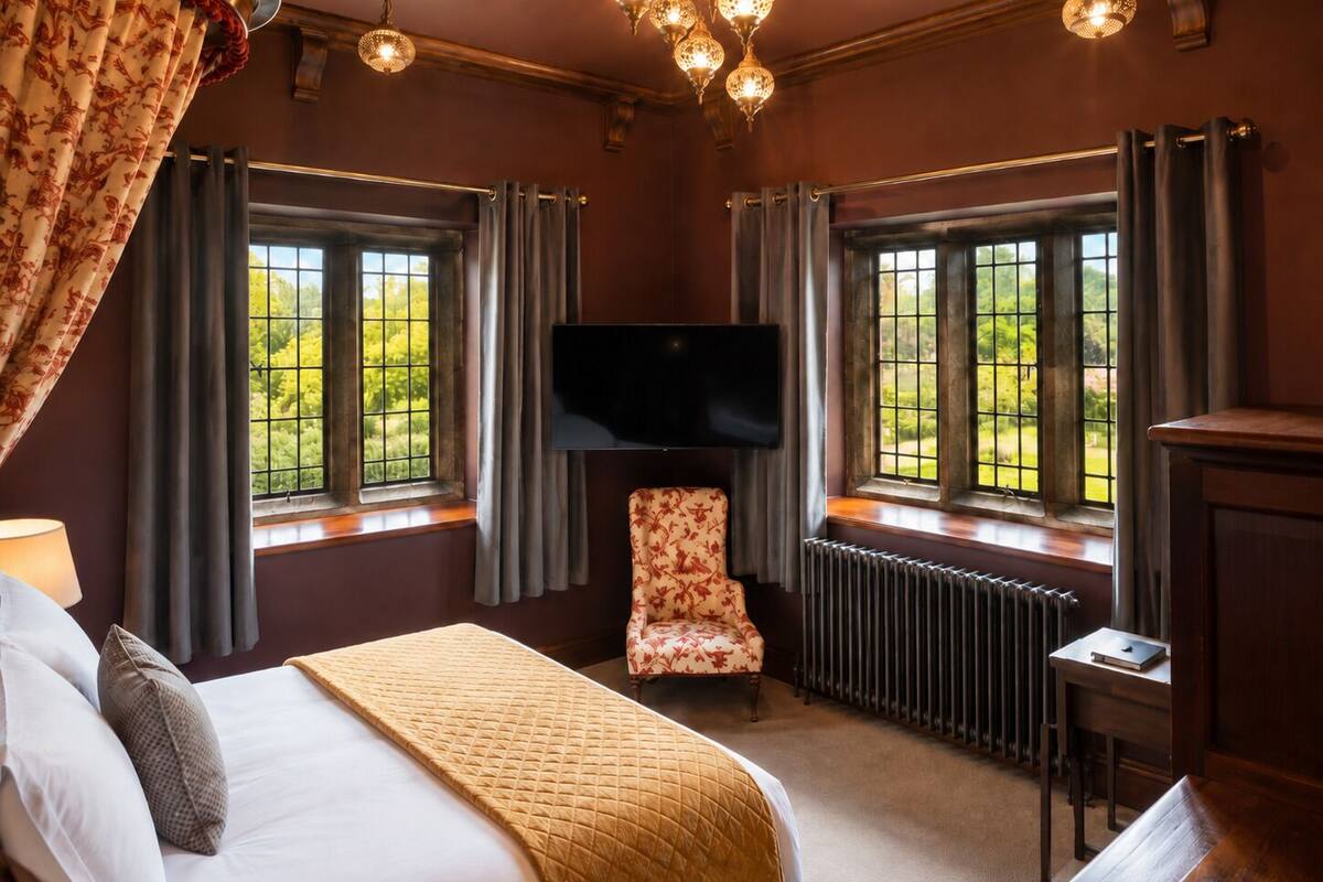 A bedroom with a bed, a chair, a TV, and windows with a view of greenery.
