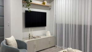 Ein modernes Wohnzimmer mit einem an der Wand montierten Flachbildfernseher, einem grauen Sofa, einem Marmortisch und transparenten Vorhängen.