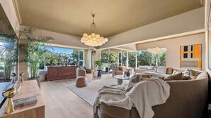 A spacious living room with a large sofa, a coffee table, and a dining area. There are large windows providing natural light and a view of greenery outside. - Spacious Kinvara Villa with AC, Gym, Pool & Mountain Views (Cape Town)