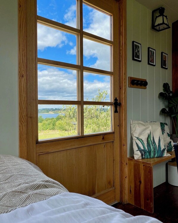 A room with a large window, a bed with patterned bedding, a wooden door, and a wall-mounted shelf with decorative pillows. - Shepherd Hut Rutland Water (Oakham)