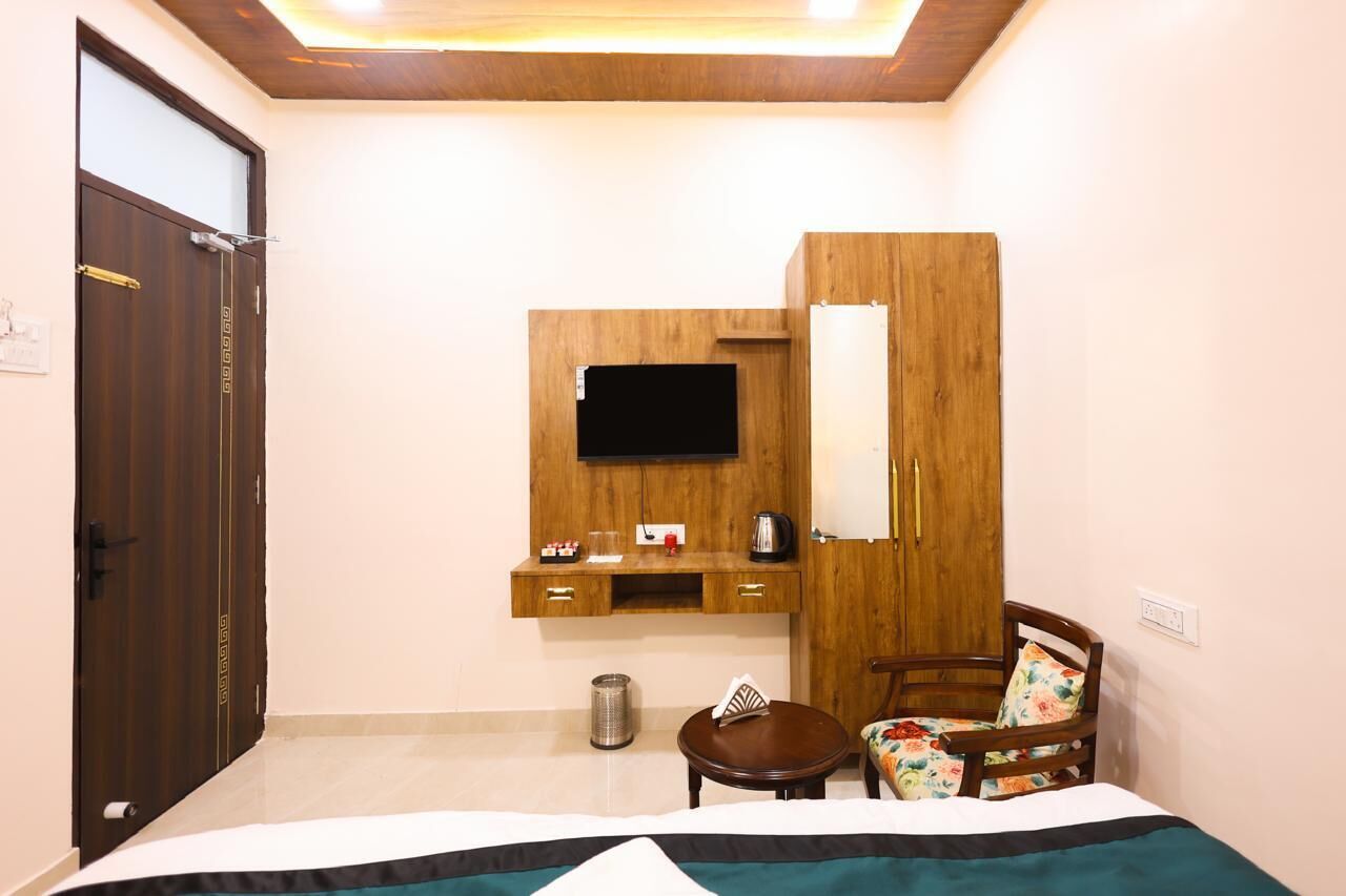 A hotel room with a bed, a wooden wardrobe, a small table, a chair, a TV mounted on the wall, and a trash bin.