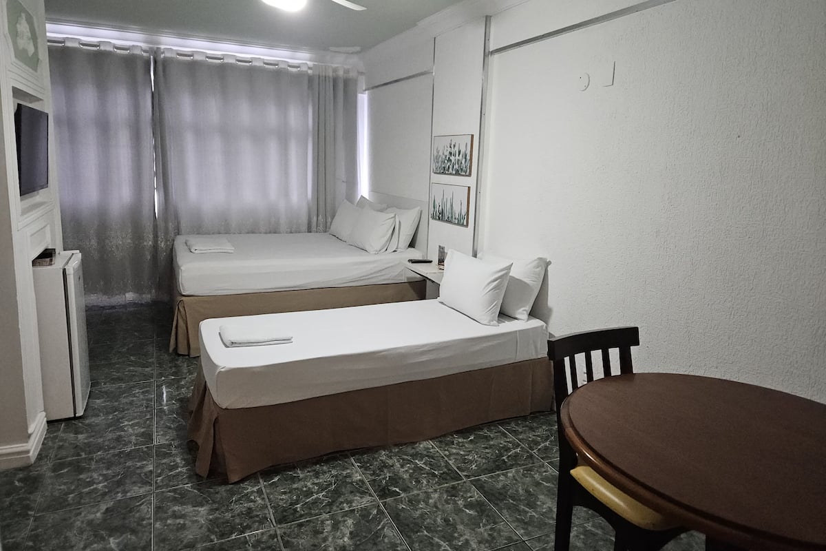 Quarto de hotel com duas camas, uma escrivaninha, uma cadeira, um ventilador de teto e uma televisão.