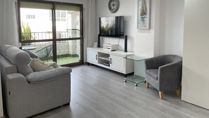 A modern living room with a grey sofa, a glass coffee table, a grey armchair, a white entertainment unit with a flat-screen TV, and a balcony with patterned tiles.