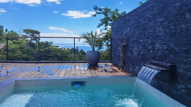 A rooftop pool with a waterfall, surrounded by a wooden deck and a stone wall, overlooking a lush landscape.