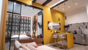 Ein modernes Hotelzimmer mit Bett, einer kleinen Küchenzeile, einem Spiegel und Blick auf eine Stadtlandschaft durch ein großes Fenster.