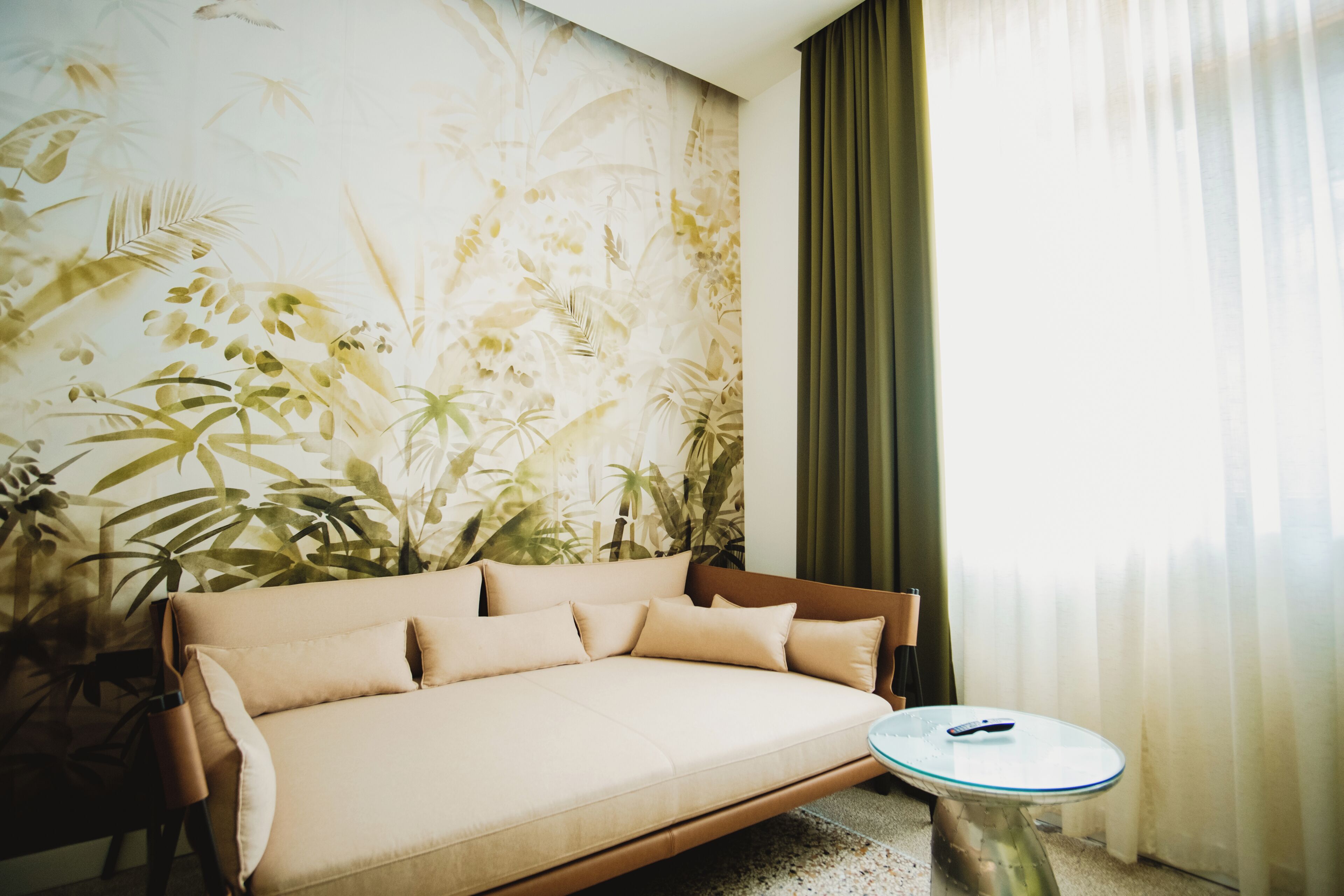 A room with a sofa, a round table, and a wall with a tropical plant mural.