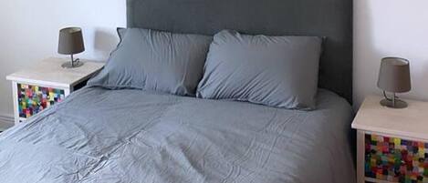 A bed with a grey headboard and pillows, two bedside tables with lamps, and a colorful storage unit.