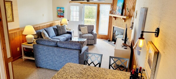 A cozy living room with a blue sofa, a patterned armchair, and a coffee table. There is a fireplace, a television, and a dining area with chairs. - Tranquil 2-bedroom condo in Duck with WiFi, AC. Enjoy your stay (Duck)