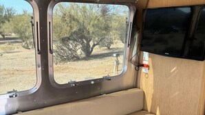 A campervan interior with a bench seating area, a mounted TV, and windows offering a view of the outdoors.