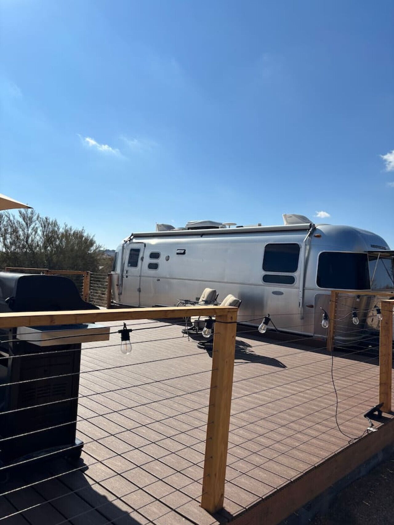 A stationary RV with a covered front area, a grill, and a table with chairs, set on a tiled deck with a clear sky in the background.