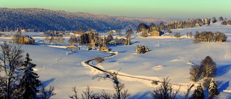A snow-covered landscape with a winding road, trees, and distant buildings.