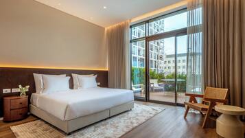 A modern hotel room with a large bed, a wooden nightstand, a round side table, and a view of the outdoors through a glass door.