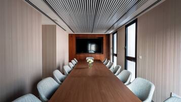 Una sala de conferencias con una larga mesa de madera, sillas tapizadas en gris, un centro de mesa floral y un televisor de pantalla plana grande en la pared.