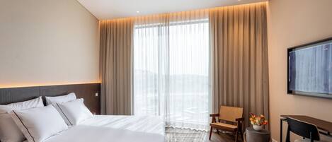 A modern hotel room with a large bed, a desk, a chair, and a TV mounted on the wall.