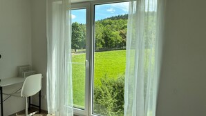 Une pièce comprenant un lit, un bureau et une chaise. Une grande fenêtre offre une vue sur un champ verdoyant et des arbres.