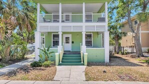 A two-story house with a green exterior, white trim, and a front porch with steps leading to a door. - Cordova Palms A - Duplex by Castillo (St. Augustine)