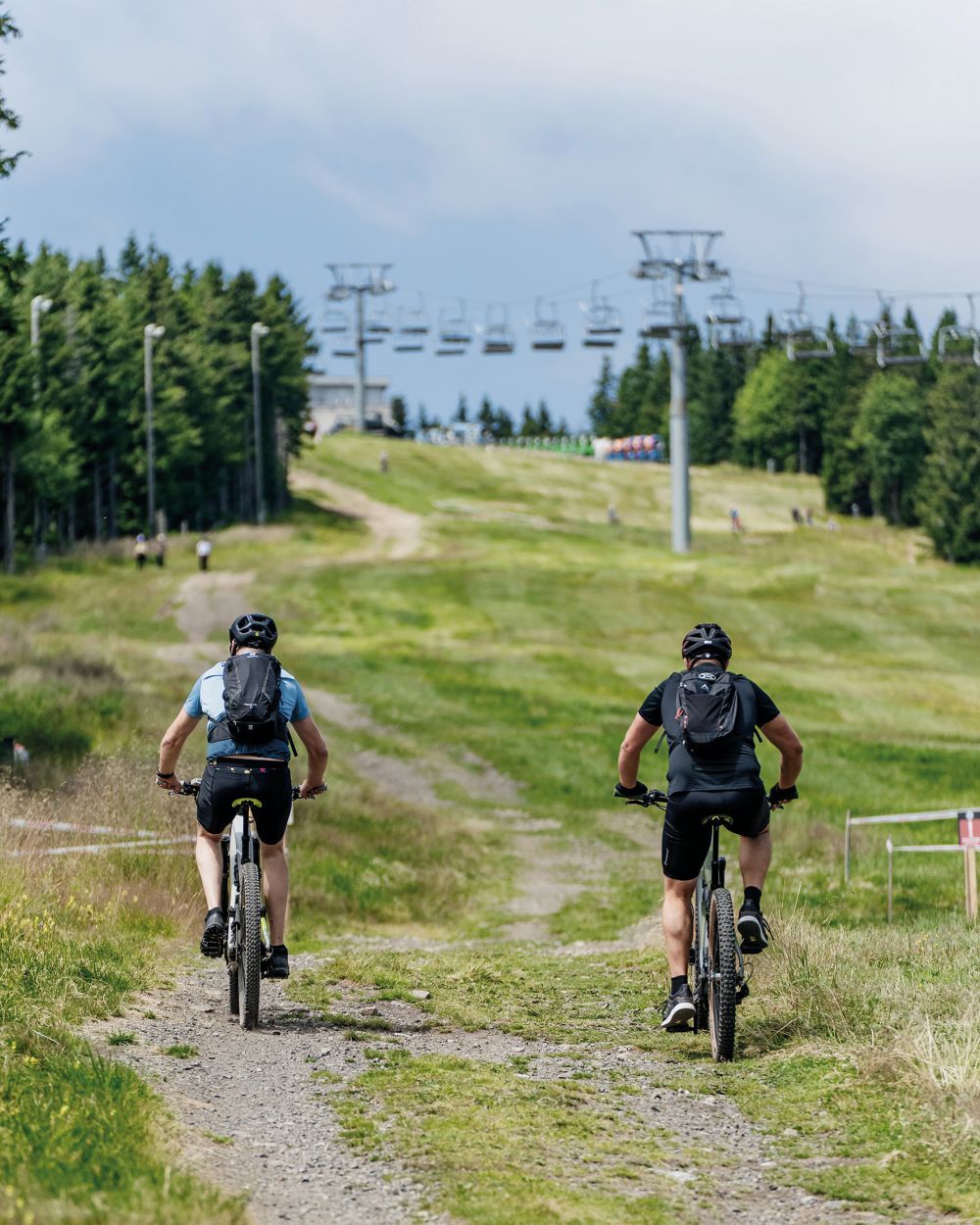 Dois ciclistas numa trilha de terra com um teleférico ao fundo.