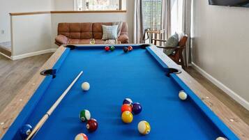 A pool table with balls, a pool cue, and a sofa in a room with a window.