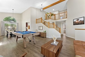 A spacious living area with a pool table, a staircase with wooden railings, and a dining area with natural light.