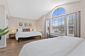 A bedroom with a large bed, a nightstand, a window with blinds, and a view of the outside.