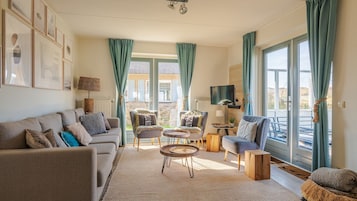 Una sala de estar con un sofá, dos sillones, una mesa de centro y un televisor. Hay cortinas, una lámpara y vistas al exterior.