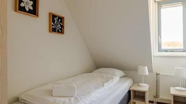 Quarto pequeno com cama de solteiro, teto inclinado, janela com vista e duas molduras de quadros na parede.