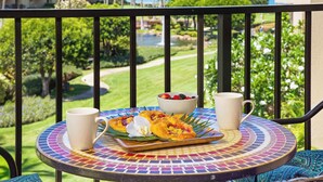 Ein Tisch mit einer farbenfrohen Mosaik-Tischplatte, eine weiße Tasse, eine Obstschale und ein Teller mit Essen, vor einer Balkonbrüstung mit Blick auf einen Garten und Gebäude.