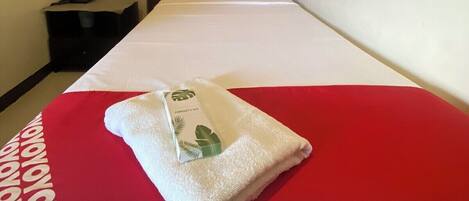 A hotel room with a bed, a red and white bedspread, a white pillow, a red pillow, a white towel, a small nightstand, a phone, and a wall-mounted air conditioner.