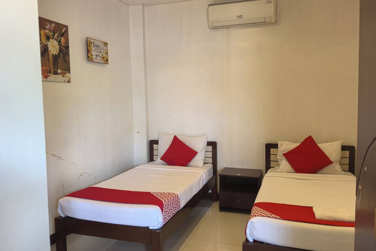 Une petite chambre propre, avec deux lits simples, chacun agrémenté d’un oreiller rouge et de draps blancs. On y trouve une petite table de chevet avec une lampe, un climatiseur fixé au mur et un tableau encadré accroché au mur.