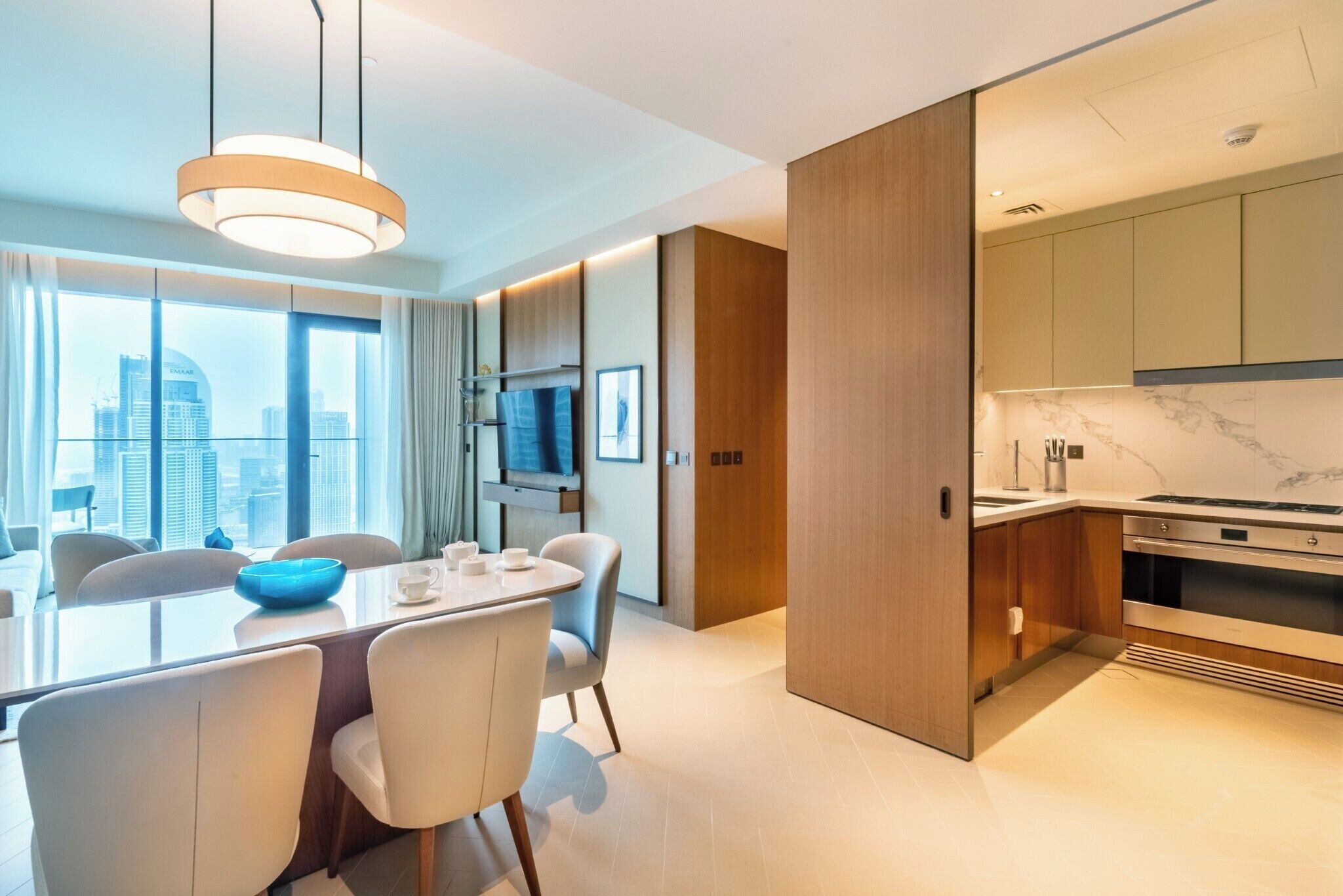 A modern hotel room with a dining area, a kitchen, and a large window offering a city view.