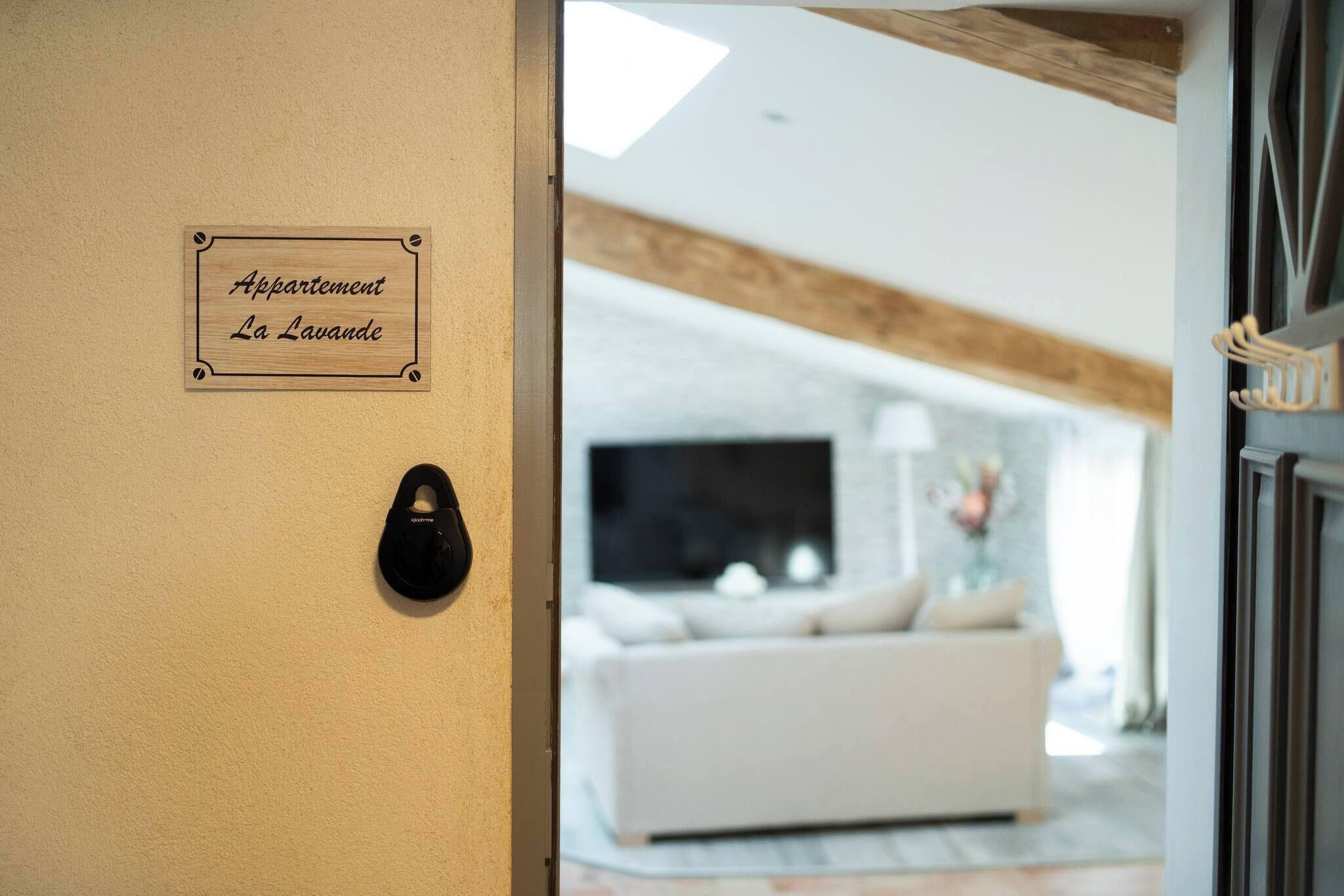 Schild mit der Aufschrift 'Appartement La Lavande' an einer beigen Wand, ein schwarzer Schuh auf dem Boden und ein Wohnzimmer mit weißem Sofa und Fernseher im Hintergrund.