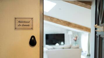 Schild mit der Aufschrift 'Appartement La Lavande' an einer beigen Wand, ein schwarzer Schuh auf dem Boden und ein Wohnzimmer mit weißem Sofa und Fernseher im Hintergrund.